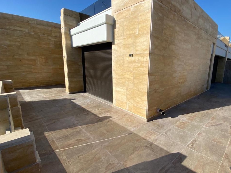Exterior view of a beige stone building with a closed black roller shutter and a white canopy, set on a tiled patio under a clear blue sky.