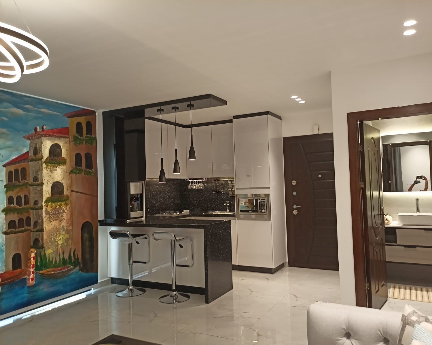 ec4099b4-f17e-431e-a6ac-2a88021cd36b Modern kitchen with black-and-white cabinetry, a speckled black island, two chrome bar stools, and hanging pendant lights; a colorful mural of a Mediterranean building runs along the left wall.