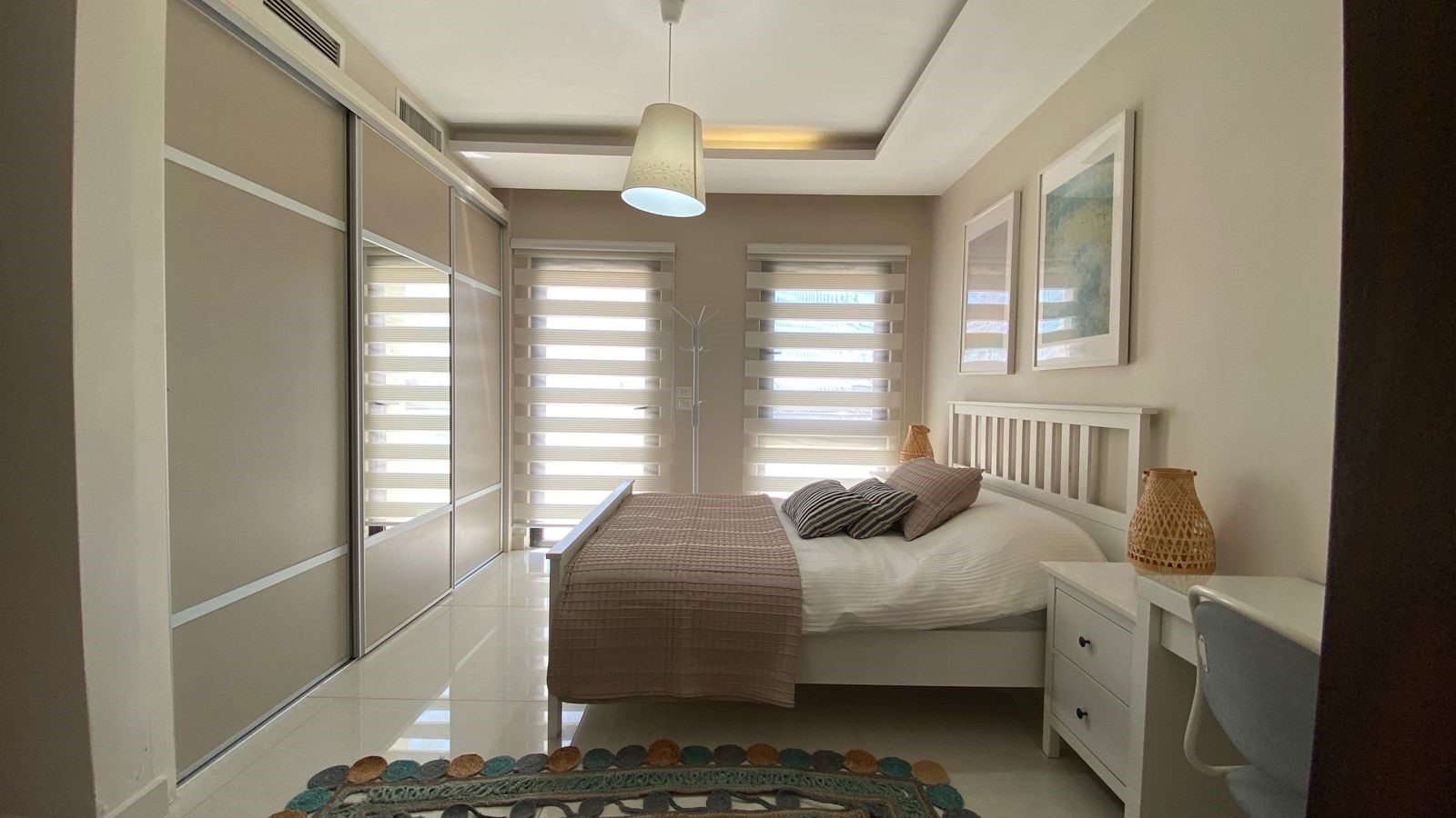ecd72830-d96c-4e96-a8c5-e84ffdf30464 Contemporary bedroom with a white slatted bed, beige bedding, two striped pillows, white nightstands, and large sliding wardrobe doors on the left.