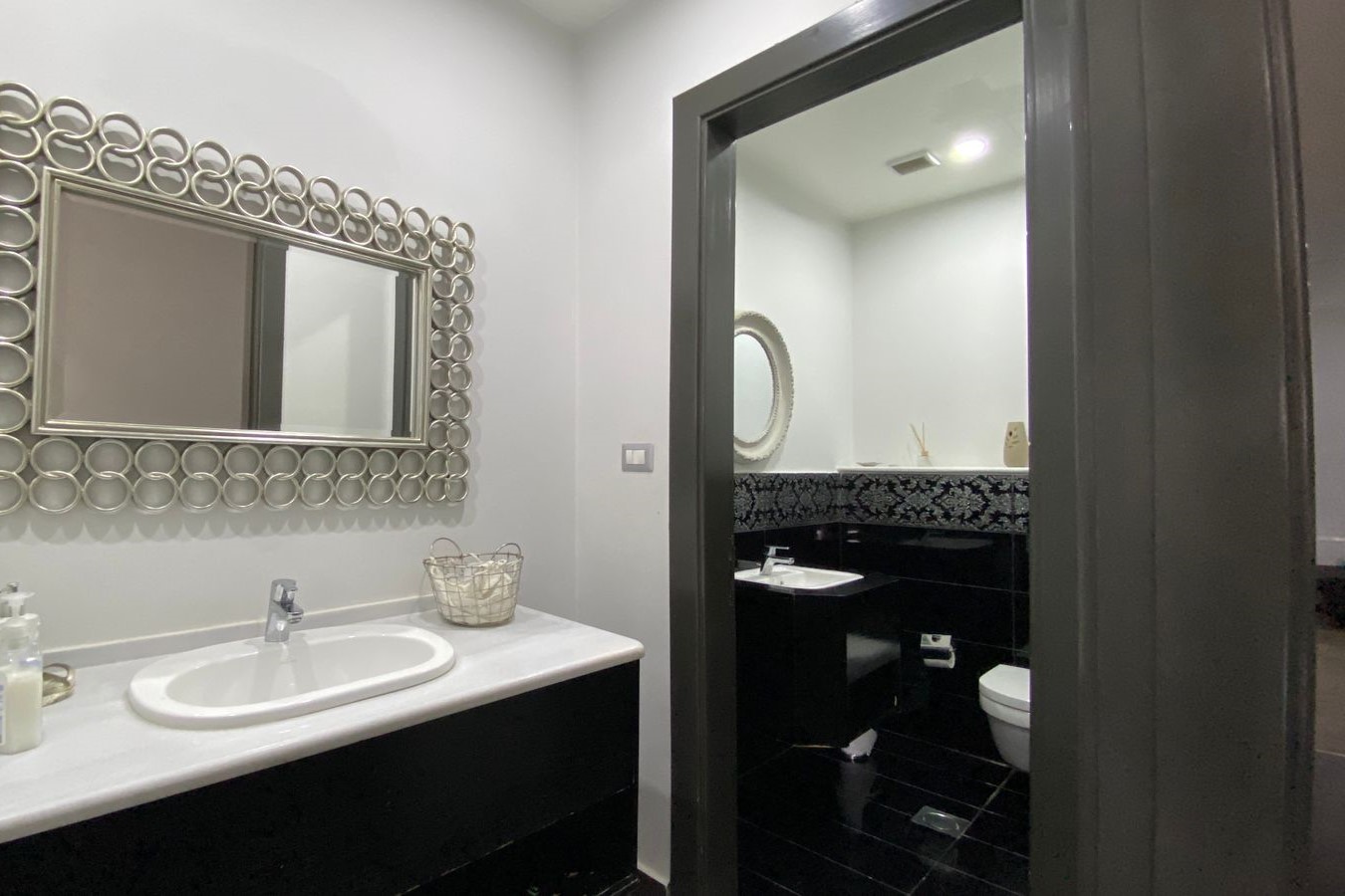 ed2556a8-56d7-45a2-83f5-48137a2a2bf5 Bathroom vanity with white countertop oval sink, chrome faucet, soap dispenser, and a decorative circular-ring framed mirror; doorway opens to a black-tiled powder room beyond.