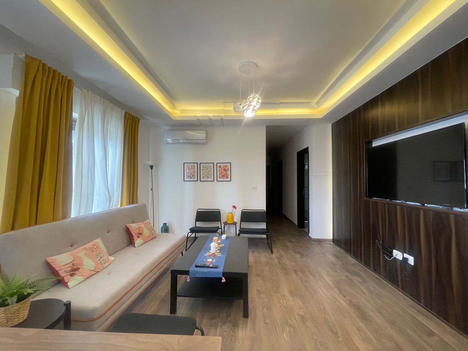 ed487201-7e13-46d4-ab30-0d19d64b0534 Modern living room with a beige sofa, mustard curtains, and a wall-mounted flat-screen TV on a dark wood panel wall visible to the right.