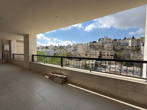 Spacious balcony with beige tile floor and black railing, overlooking a hillside city of white stone buildings under a blue sky with scattered clouds.