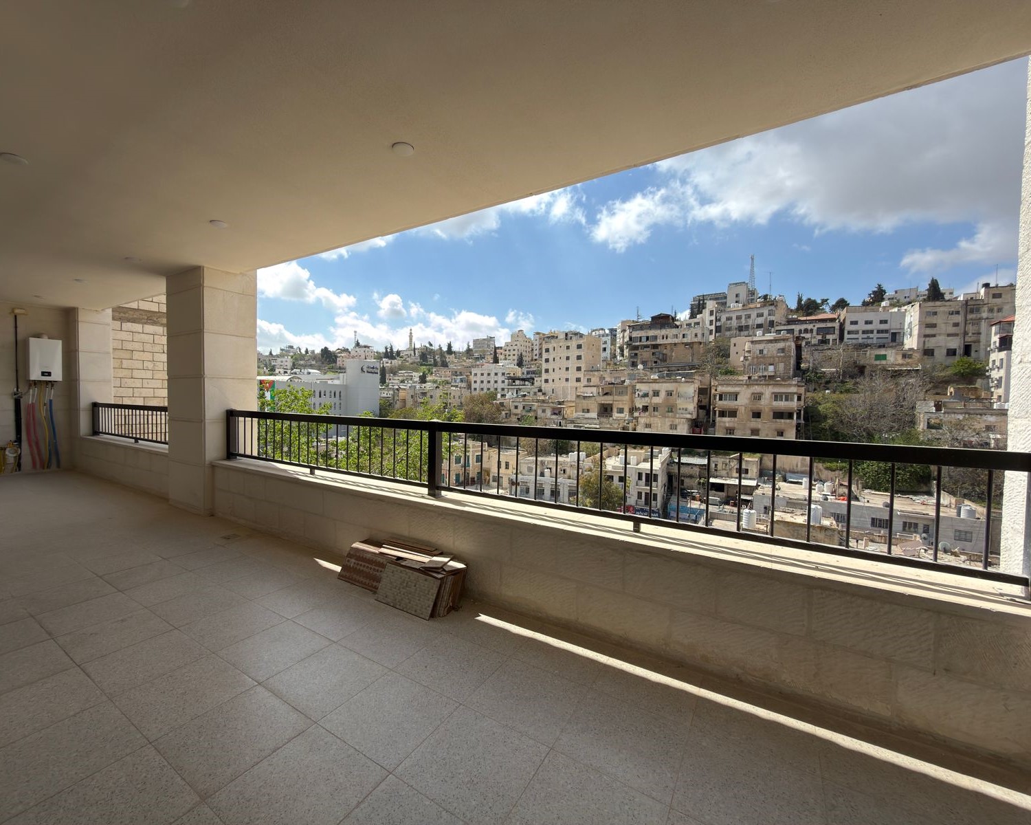 edb30c23-ccc7-4c51-8cbc-3357ca1e99c9 Spacious balcony with beige tile floor and black railing, overlooking a hillside city of white stone buildings under a blue sky with scattered clouds.