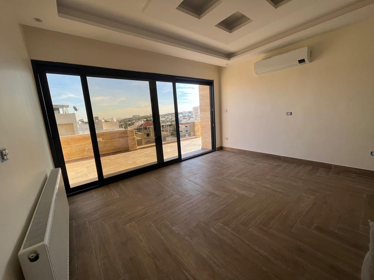 ee4646d3-2c34-4f16-ba92-c9070c806299 Empty living room with large sliding glass doors opening to a balcony and city view; wood-look tiles and a wall-mounted AC unit.