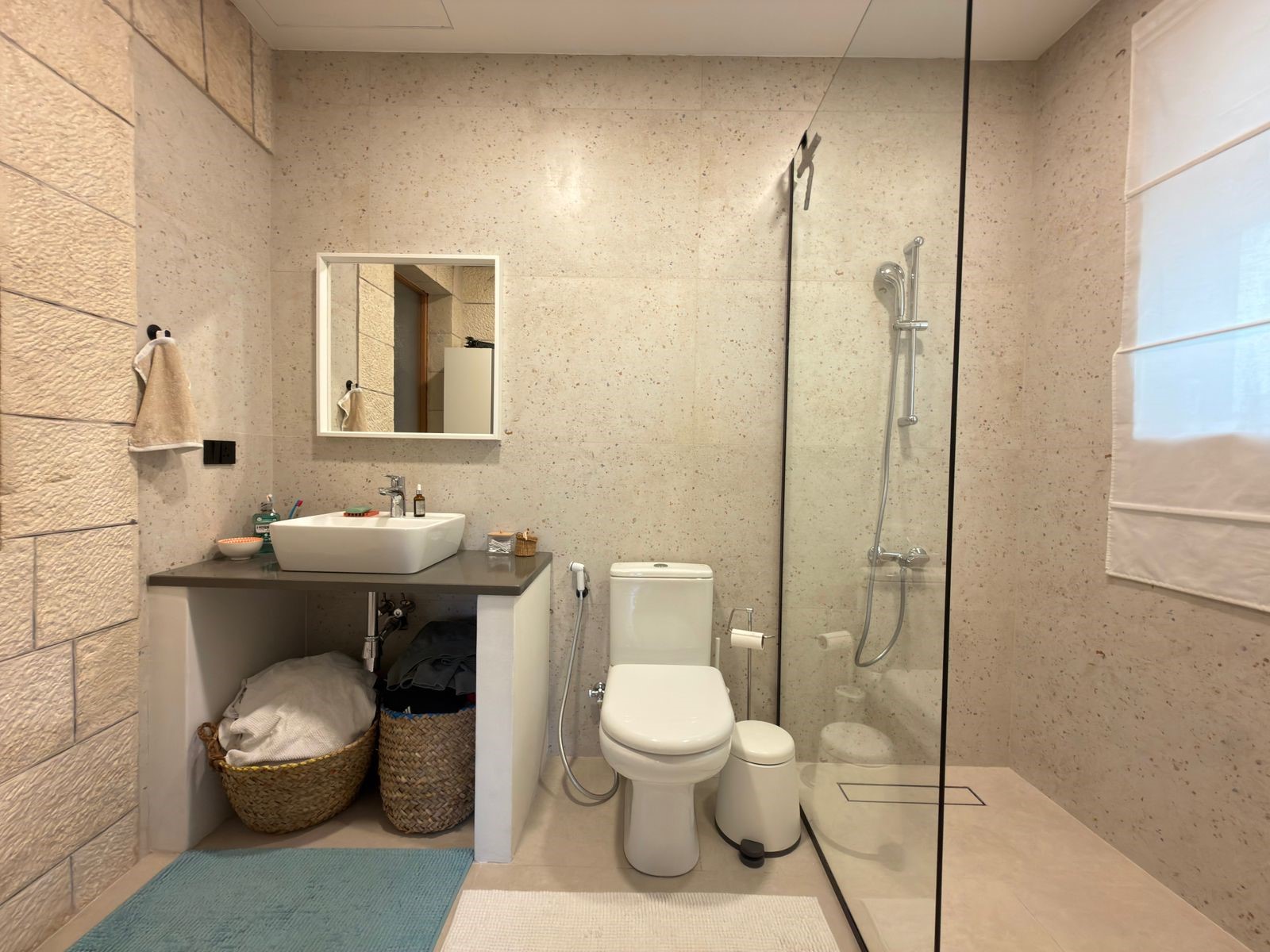 ee52619f-20e4-454a-8c2c-60cc0e7baa71 Contemporary bathroom featuring a glass-enclosed shower, white toilet, and a sink on a dark countertop with wicker baskets underneath.