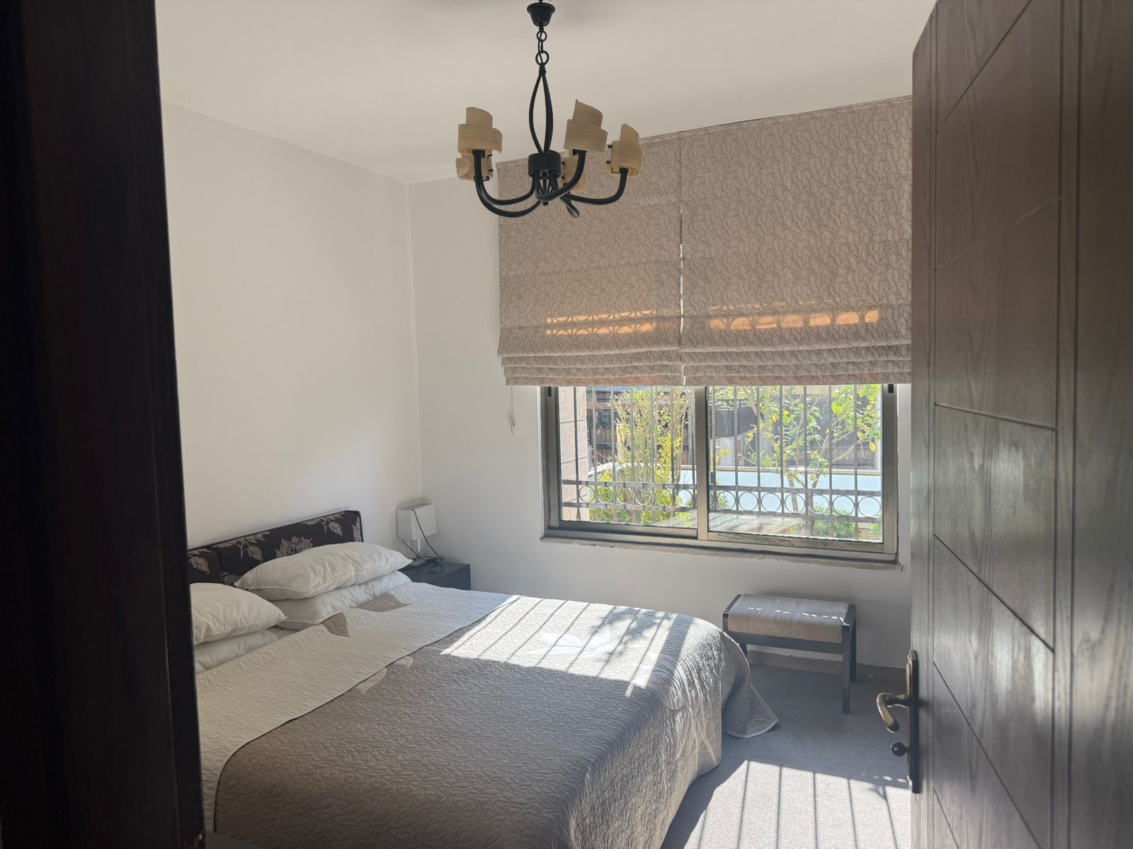 ef07bc25-498f-4dfa-a89e-17fdf40bd4f4 Bedroom with a double bed, beige quilts and white pillows; window with patterned blinds and a black chandelier overhead.