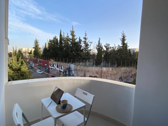 Small balcony with a white table and two folding chairs; laptop and a cup on a woven placemat, overlooking a graffiti wall and trees.