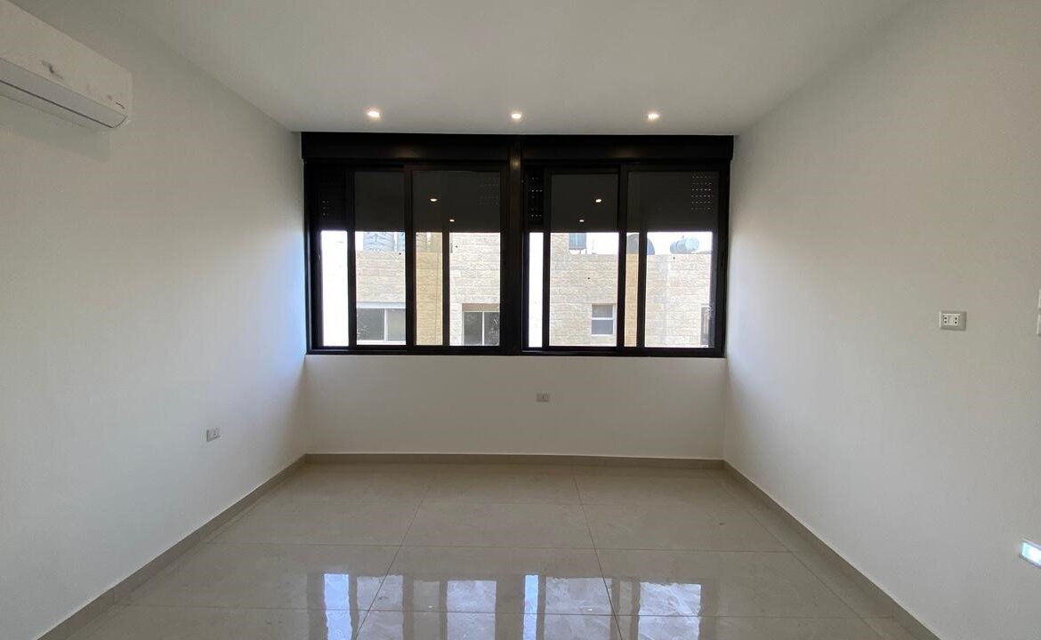 Empty white room with a large black-framed window, tiled floor, and wall-mounted air conditioner on the left.