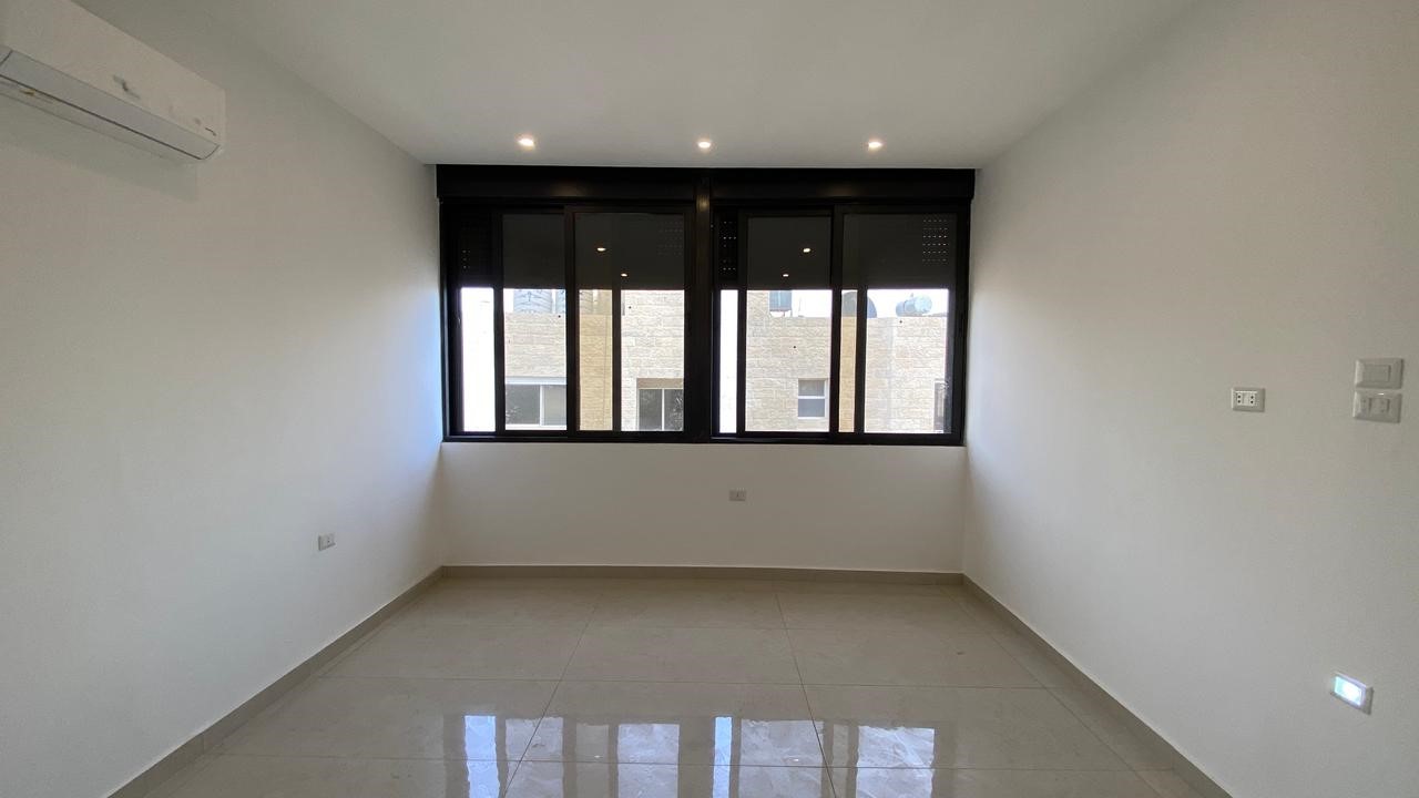 f082b86e-c16d-4401-86eb-4893b1088650 Empty white room with a large black-framed window, tiled floor, and wall-mounted air conditioner on the left.