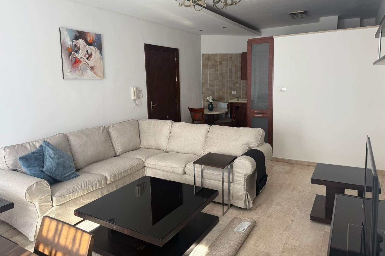f14fb669-3c5f-41de-9a56-38c15ae97dd2 Cozy living room with a light beige sectional sofa and blue pillows facing a black glass coffee table and TV stand. A dining nook and tiled wall are in the back.