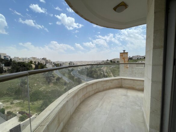 Curved balcony with glass railing overlooking a cityscape and a winding highway beneath a blue, partly cloudy sky.