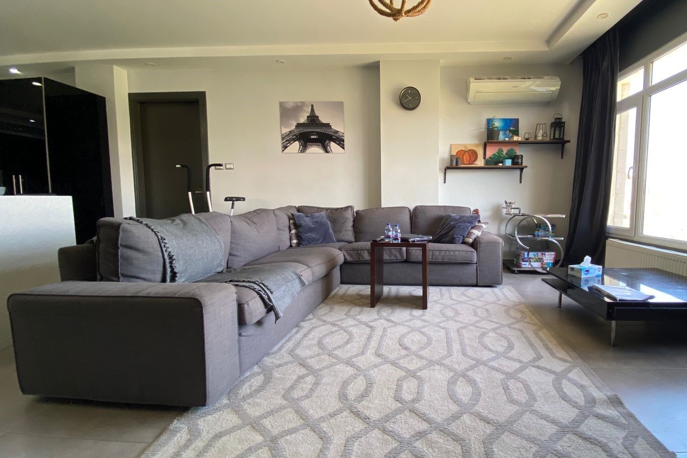 f248c324-23cc-4641-af73-00f78514c262 Modern living room with a large gray sectional sofa, patterned rug, and a glass coffee table.