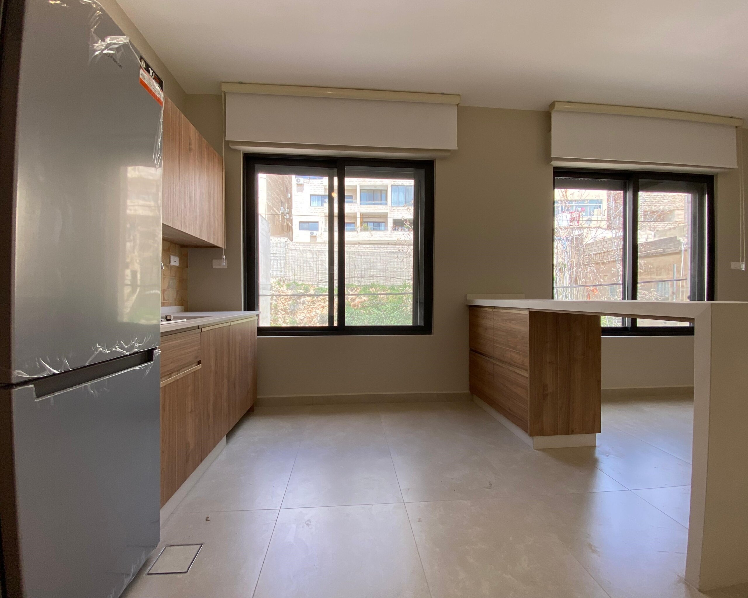 f353681b-4354-4ea4-8382-a1a433cdc593 Modern kitchen with wooden cabinets, a tall stainless steel fridge on the left, and a wooden island under a bright windowed wall.