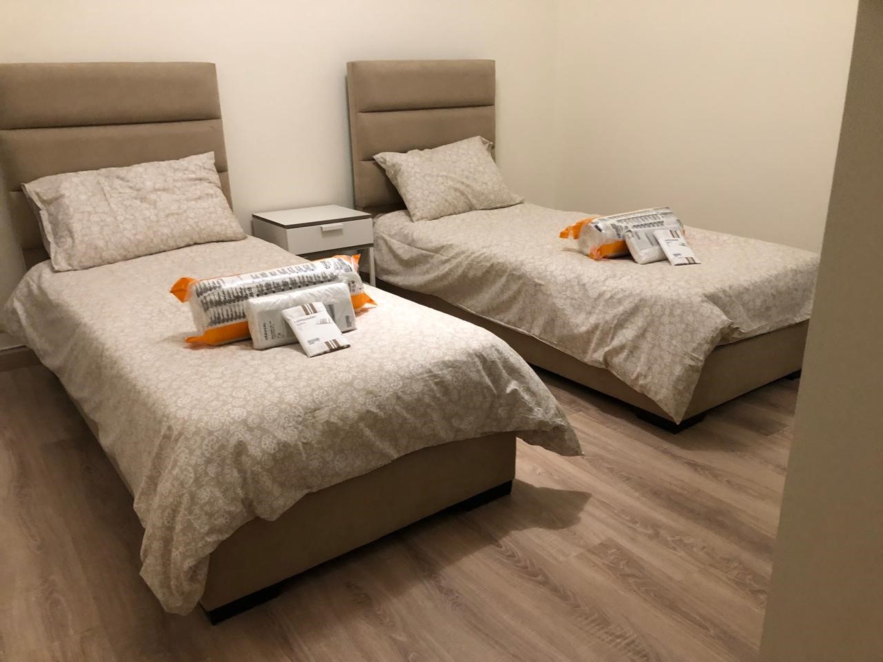 f36e783e-7e0a-4539-bf44-35e1579f6504 Two twin beds with beige upholstered headboards, a white nightstand between, and packaged linens on each bed in a neutral hotel room.