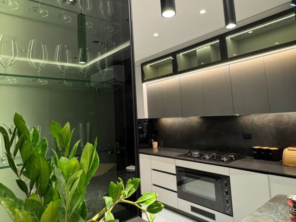 Modern kitchen with gray upper cabinets, built-in oven and gas cooktop, glass-front cabinet with wine glasses on the left, and green plants in the foreground.