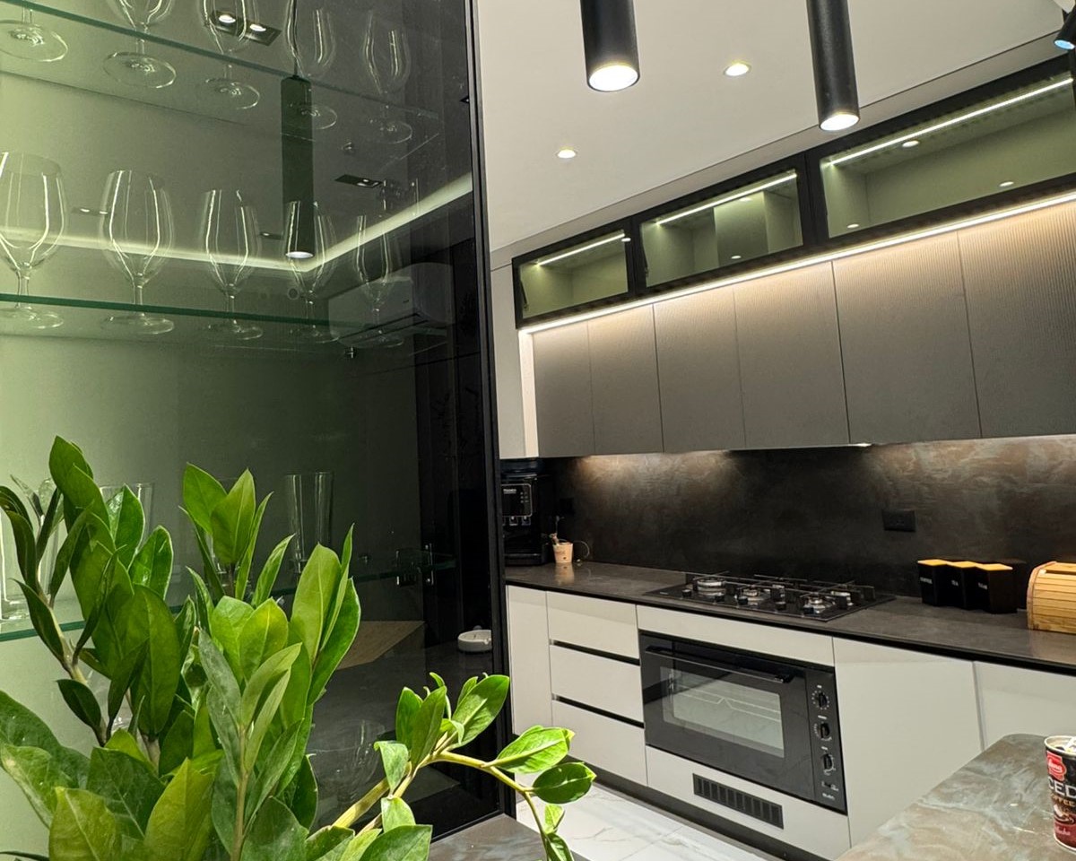 f3d6a00d-2522-4b53-9f80-5b1288ba6ff4 Modern kitchen with gray upper cabinets, built-in oven and gas cooktop, glass-front cabinet with wine glasses on the left, and green plants in the foreground.