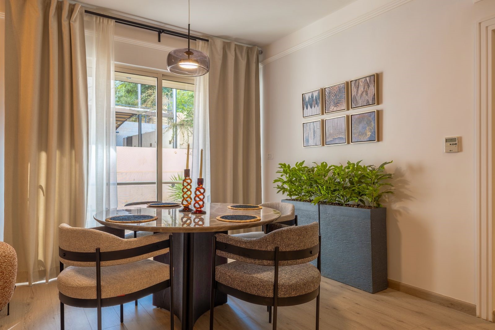 Round glass-top dining table with four upholstered chairs in a modern dining nook; plant-filled blue planter against the wall and a grid of framed artwork above it