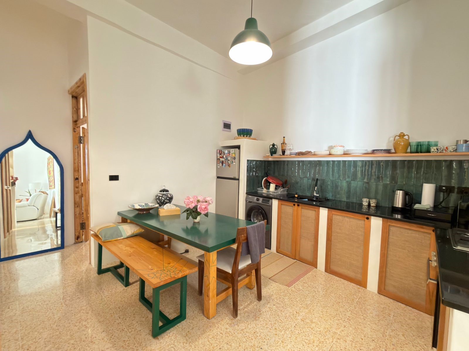 f50f7811-404e-4ba8-ac5f-bbb30ff6321f Open-plan kitchen and dining area with a green table and wooden benches, teal tiled backsplash, and shelves of dishes along the counter.