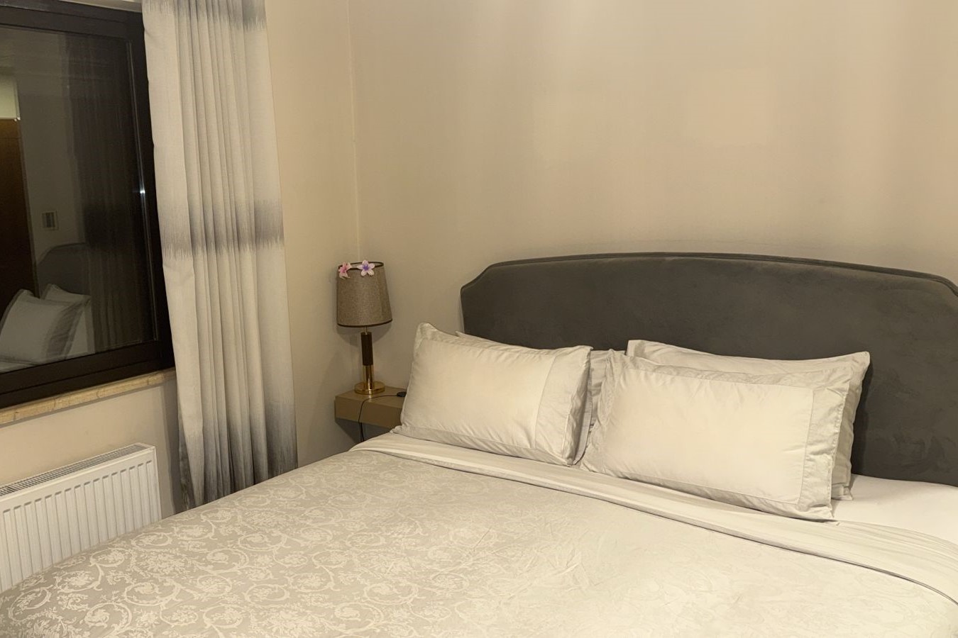 f6155db6-25eb-402b-ac5f-0492a025f4f7 1 Hotel bed with white linens and a gray headboard, beside a nightstand lamp in a room with a window and curtains.