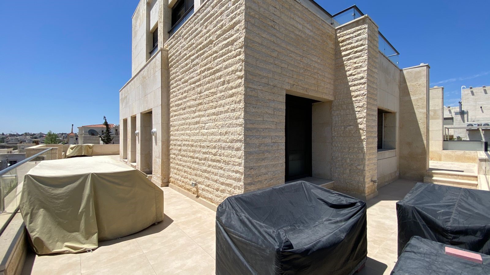 f660c85b-c8f2-40ca-a6da-1dbd215f47ff Rooftop terrace with beige stone walls and covered outdoor furniture on a sunny day.