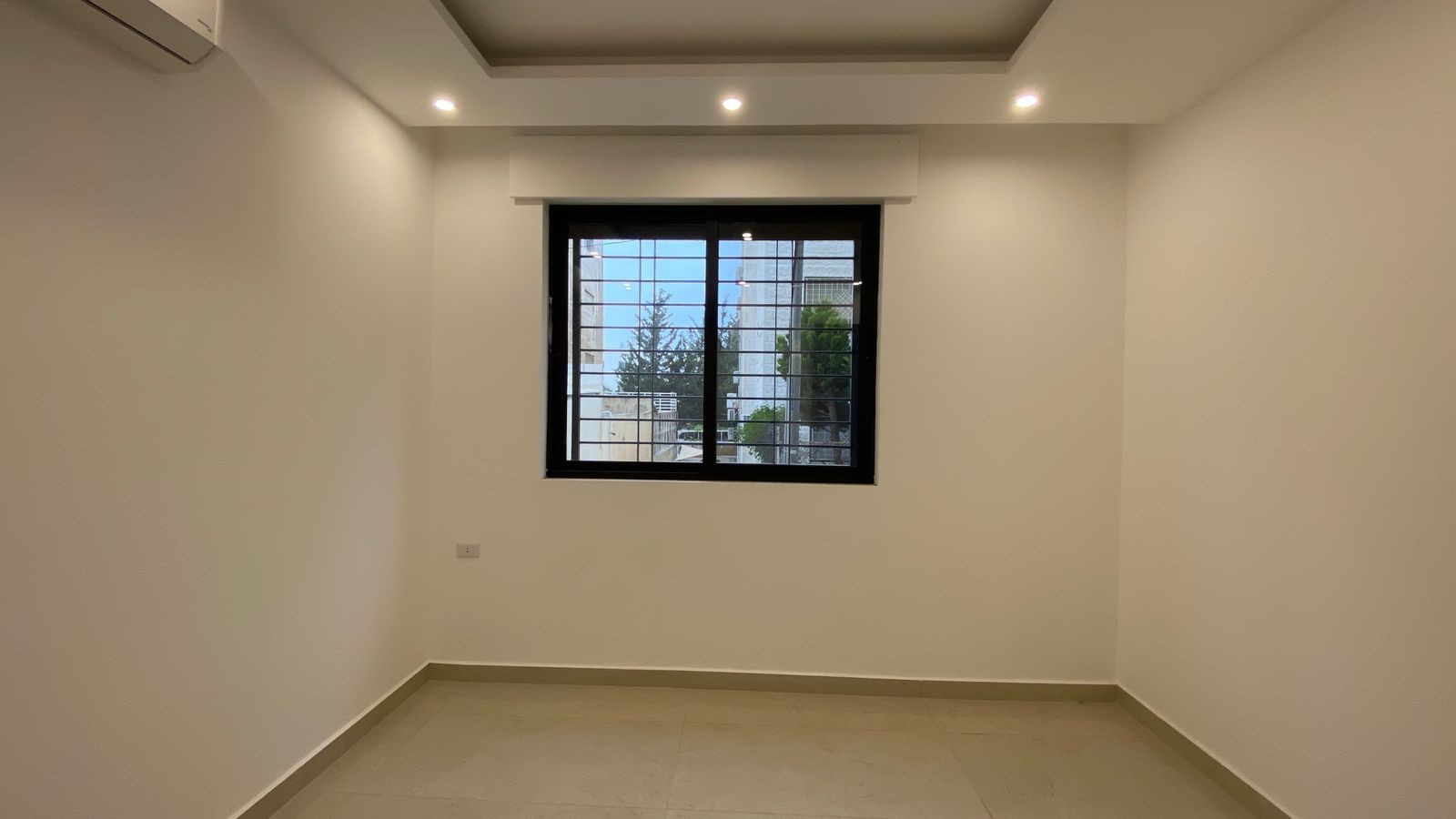 f7c11891-07ac-4f79-b935-c38c454e89d7 A small empty white room with a square recessed ceiling, a black-framed window, and tiled beige floor.
