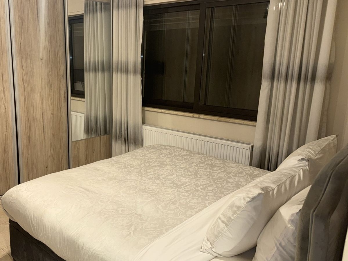 f847197b-1481-4bd6-a2bf-84bc486fb319 Modern bedroom with a large bed covered in a white patterned duvet, grey pillows, a wooden wardrobe with a mirror, and a window with light curtains.