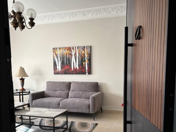 Living room view: gray sofa with pillows, marble coffee table, side lamp, and a birch-tree artwork on a beige wall near the doorway.