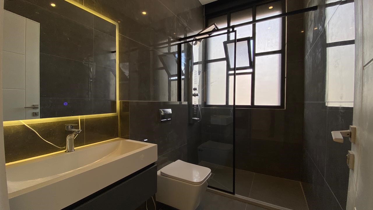fb23c59d-d00a-4616-9c73-21a666a3c572 Modern bathroom with a backlit rectangular mirror, white sink, wall-mounted faucet, wall-hung toilet, and a glass shower enclosure with a black frame; dark tiled walls and frosted windows.