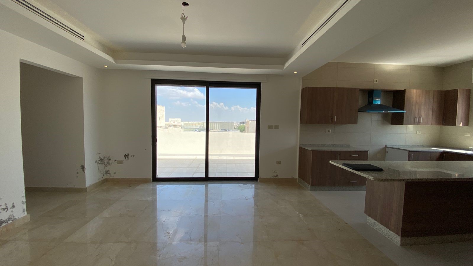 fbd8a4f0-b7bd-4d1f-bf68-6b3c2fb22f95 Open-plan living area with a sliding glass door to a balcony, leading to a distant city view; adjacent modern kitchen with wooden cabinets and granite island; glossy tiled floor and a bare bulb hanging from the ceiling.