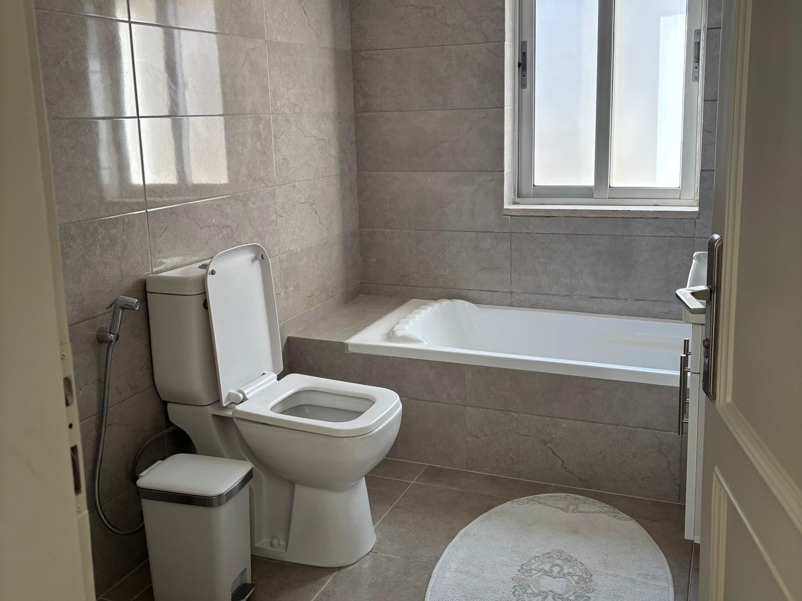 fbfc6810-6e98-42c2-87b1-f875c08584b2 Modern bathroom with gray tiled walls, a white toilet with lid up, a handheld bidet spray, and a built-in bathtub beneath a window. A small white trash can sits beside the toilet, and a white oval rug lies on the floor.