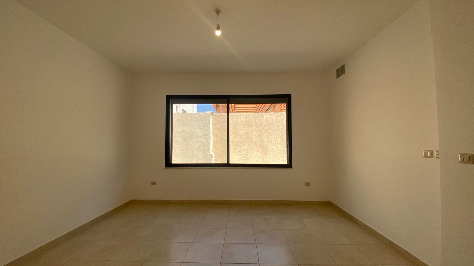 fc4dd216-7e85-4352-b7fc-1cc97c563816 Empty rectangular room with off-white walls, beige tiled floor, and a large window showing a beige exterior wall outside.
