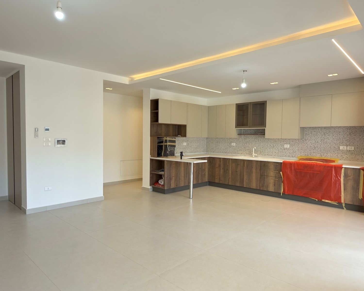 fc92135f-ac66-4335-83be-fb359d442eef Open-plan kitchen with beige upper cabinets, dark wood lower cabinets, grey mosaic backsplash, and red protective wrap on the island area (under construction).