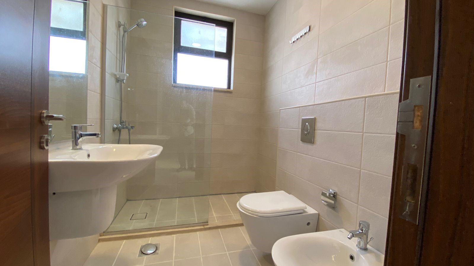 fdbe7315-bfee-43eb-88cd-5106b07b5a05 Contemporary bathroom with a glass-enclosed shower, pedestal sink, wall-mounted toilet and bidet, and beige tiled walls. A small window sits above the shower.