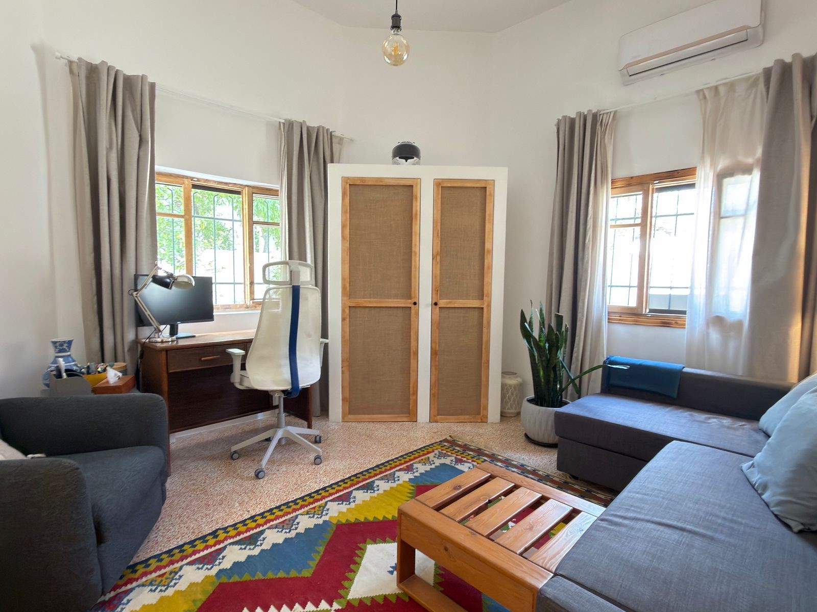 fe04373d-4bc3-4b45-bbdd-bb8a2b848d8e Bright living room used as a workspace: white ergonomic chair at a wooden desk with monitor, near a tall wardrobe between windows.