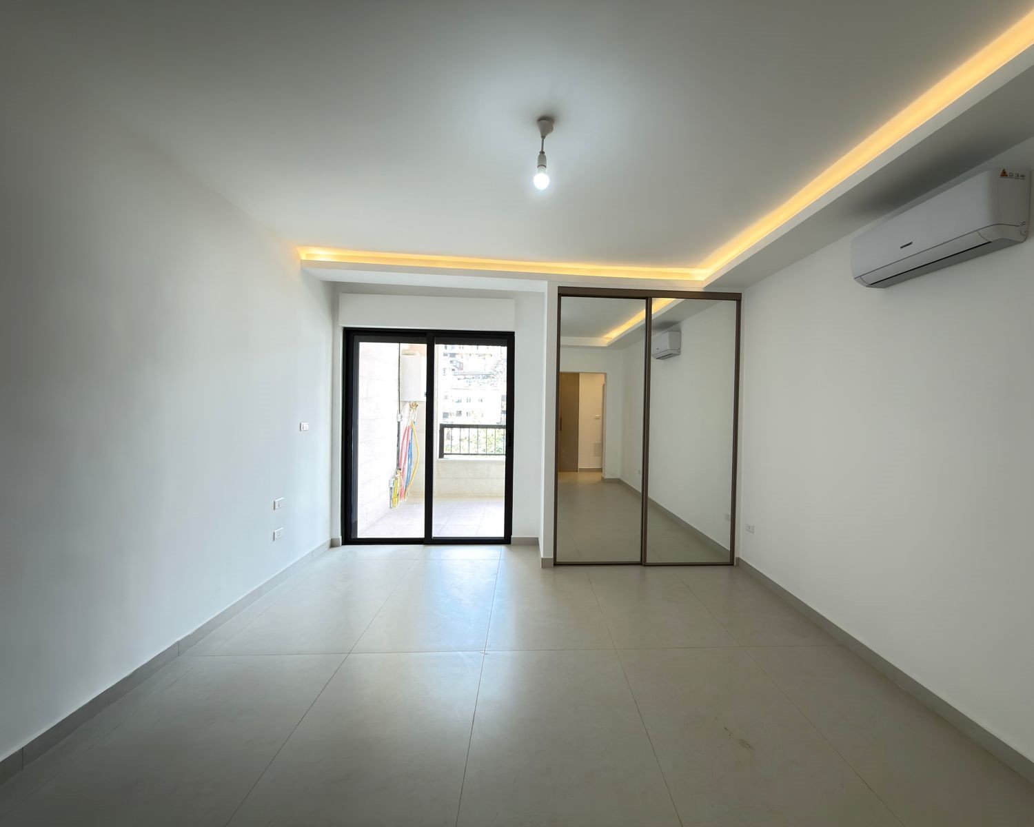 fe7123ab-eb0d-49ba-b809-4c39d5c3eb04 Empty living room with tiled floor, sliding glass door to a balcony, and a mirrored wardrobe; ceiling has warm recessed lights and a wall-mounted AC unit above.