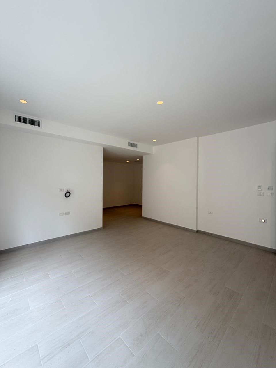 feee77a4-4a4e-4978-b5b0-3457e4ff8a67 Empty, unfurnished living room with gray tile floor, white walls, and recessed ceiling lights; doorway leads to a hallway beneath a beam.