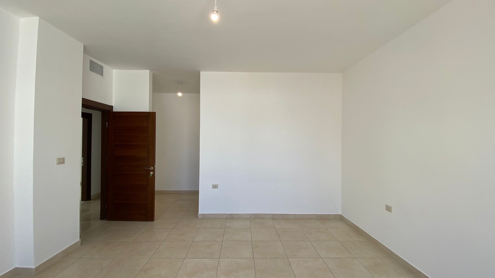ffb3d473-fe03-4dc3-8443-b35e06b1739a Empty, white-walled living space with beige tiled floor and an open wooden door leading to a hallway