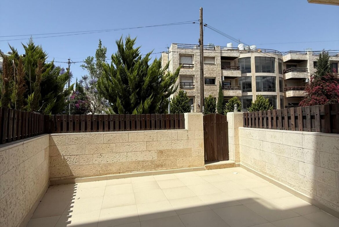 Sunny tiled courtyard with beige stone walls and a wooden fence; modern apartment building in the background under a clear blue sky.