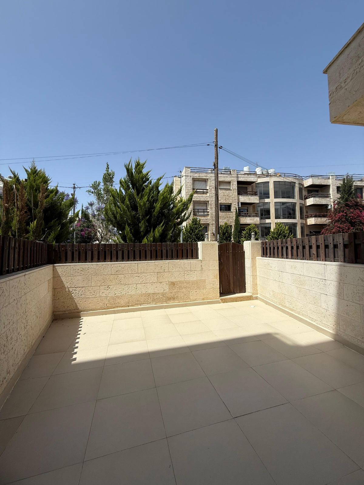 Sunny tiled courtyard with beige stone walls and a wooden fence; modern apartment building in the background under a clear blue sky.