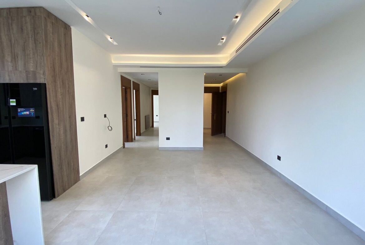 Modern open-plan kitchen and hallway with wood accent cabinetry, black fridge, and white island; bright, empty space awaiting decor.