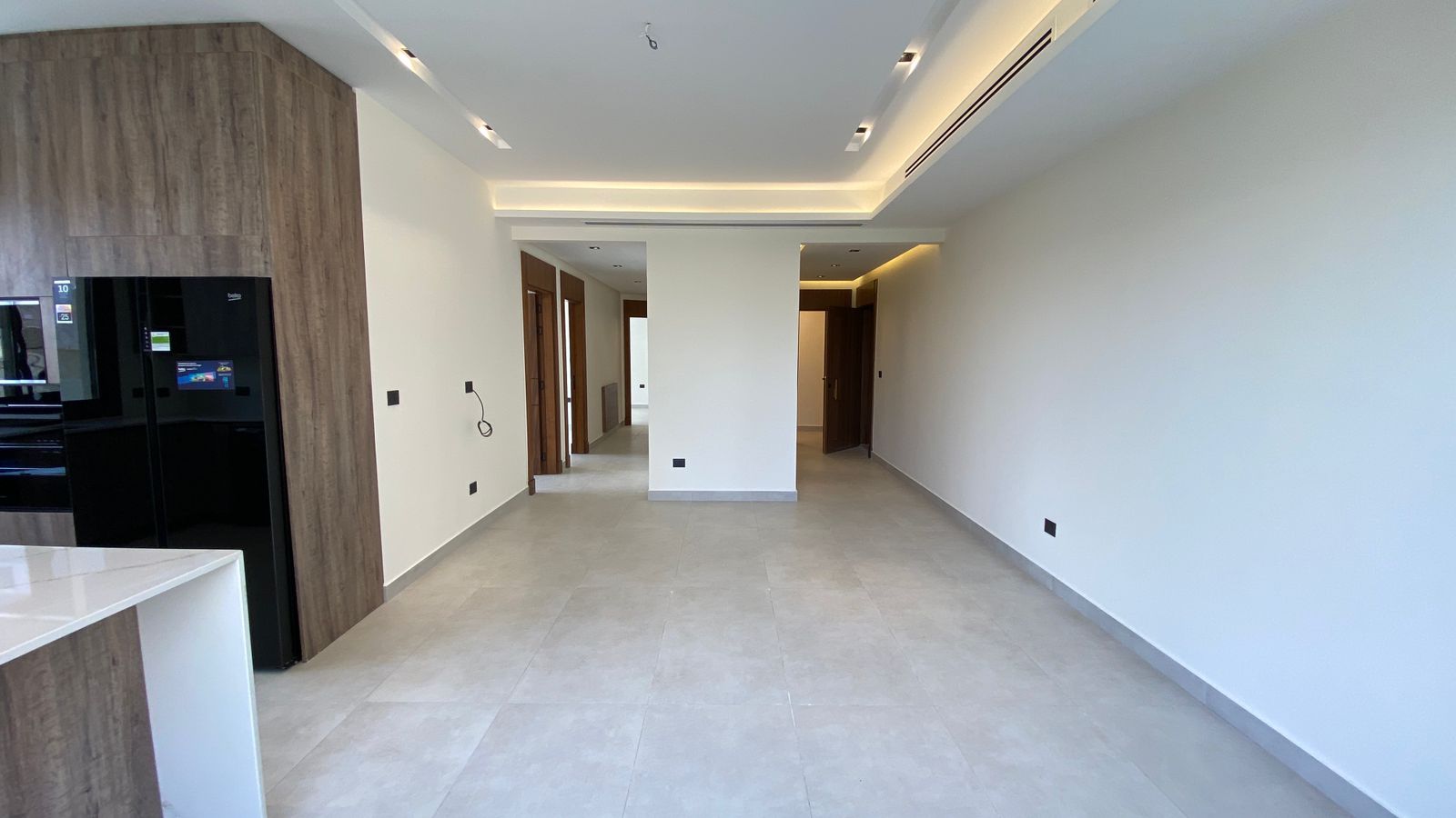 00d222d6-9071-4f0d-999f-15818a41ddb2 Modern open-plan kitchen and hallway with wood accent cabinetry, black fridge, and white island; bright, empty space awaiting decor.