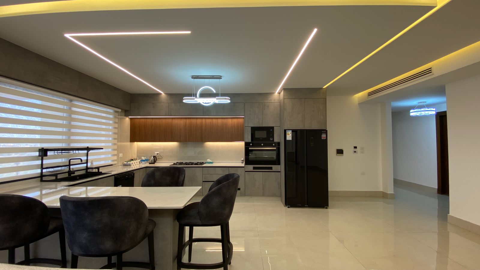 010ed782-de57-40b7-bec8-ac5a82b48061 Modern open-plan kitchen with marble island, gray cabinetry, black stools, built-in oven/m microwave, and a black side-by-side fridge. Elegant ceiling lighting adds a futuristic feel.