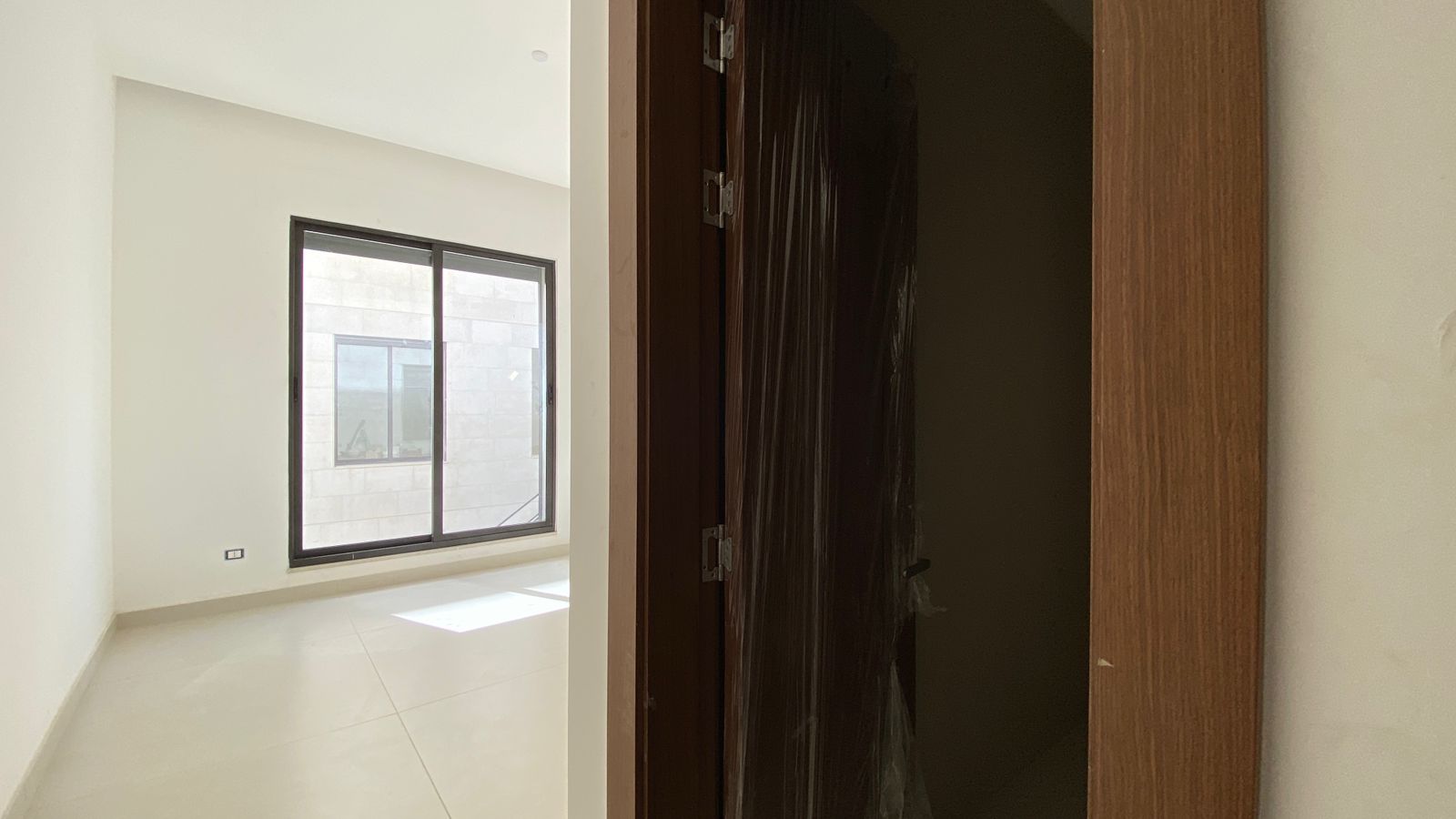 012039b1-57e6-46f1-b364-b90aca1a595e Bright empty room with a large window letting in sunlight; doorway on the right with a dark plastic-wrapped door.