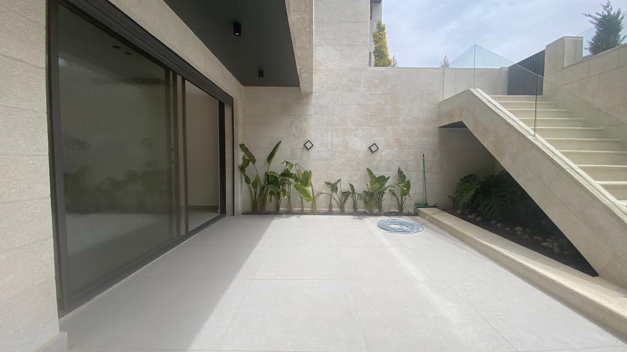 01c16396-c0c5-40d4-820b-a376c9f5a245 Modern beige stone patio with large sliding glass doors, a row of tropical plants, and a concrete staircase with glass railing to the right.