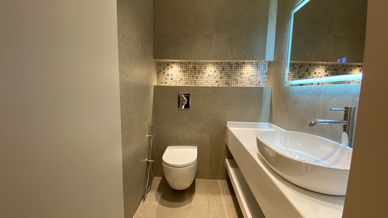 021c768b-e10c-4453-a3ea-e96649b62cbc Modern gray bathroom with a wall-mounted toilet, white vessel sink, chrome faucet, and a mosaic tile backsplash under a backlit mirror.