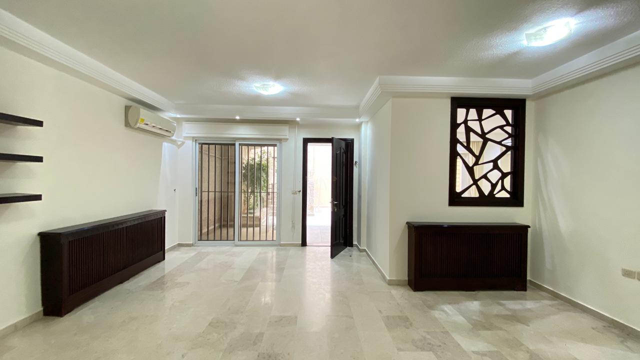 028aa8ef-0867-49de-a9f2-c41092cffd16 Bright living room with light walls and marble floors, featuring dark wood cabinets along the left wall and a decorative lattice window on the right.