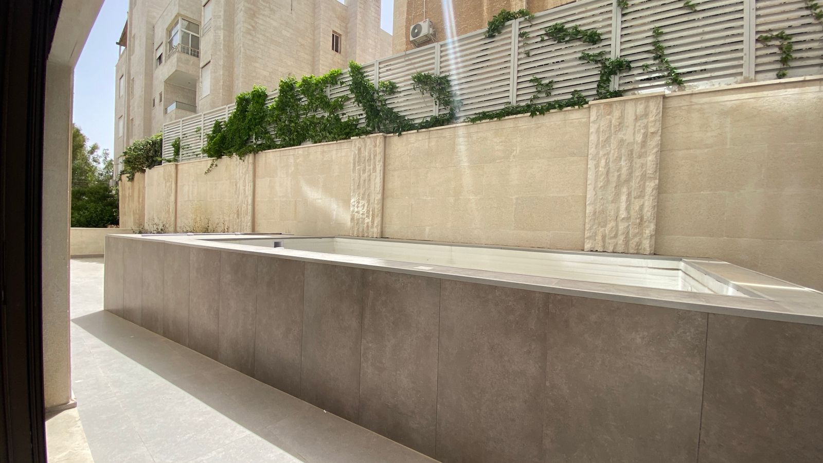 02c896df-63ac-4f45-810a-9927e47e0810 Wide view of a minimalist beige courtyard with a sunken rectangular pool bordered by gray stone panels.