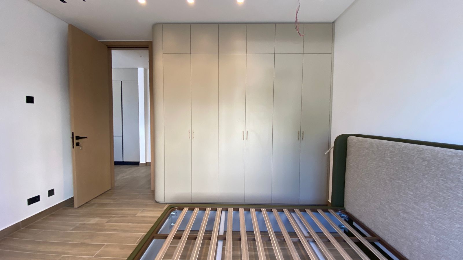0301348d-ac57-46c3-8e01-ad2f1bf5149c Bedroom with a tall white built-in wardrobe, an open wooden door to a hallway, and a metal bed frame with a green upholstered headboard at the right; ceiling light wires visible.