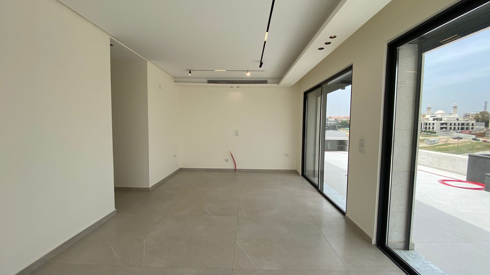 0318b8fb-253f-449a-9ba0-581cd1d5934c 1 Empty modern living room with large glass sliding doors to a balcony, gray tiled floor, and white walls; a red conduit protrudes from the wall near the center.