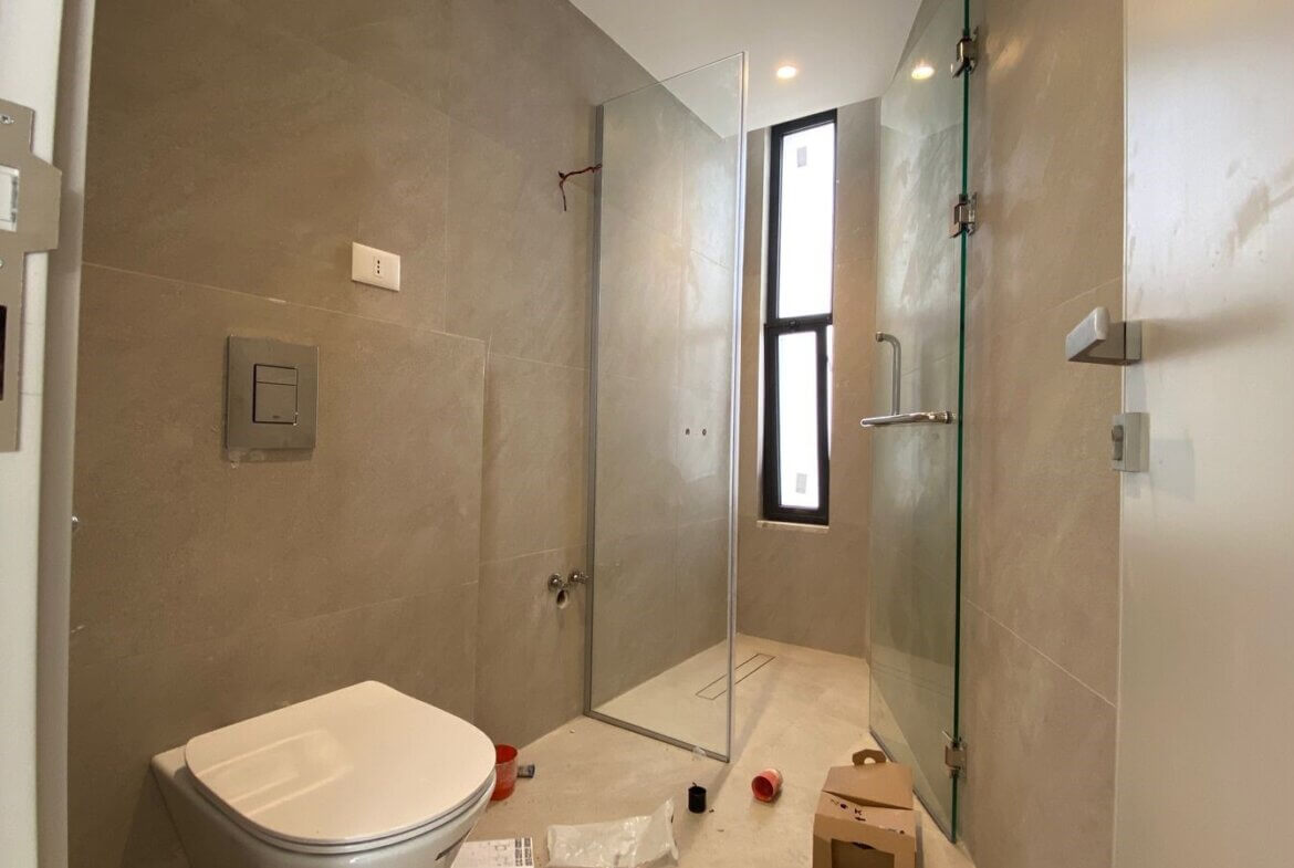 Unfinished modern bathroom featuring a wall-mounted toilet, beige tile walls, and a glass shower enclosure with a tall narrow window. Construction items and packaging debris are on the floor.